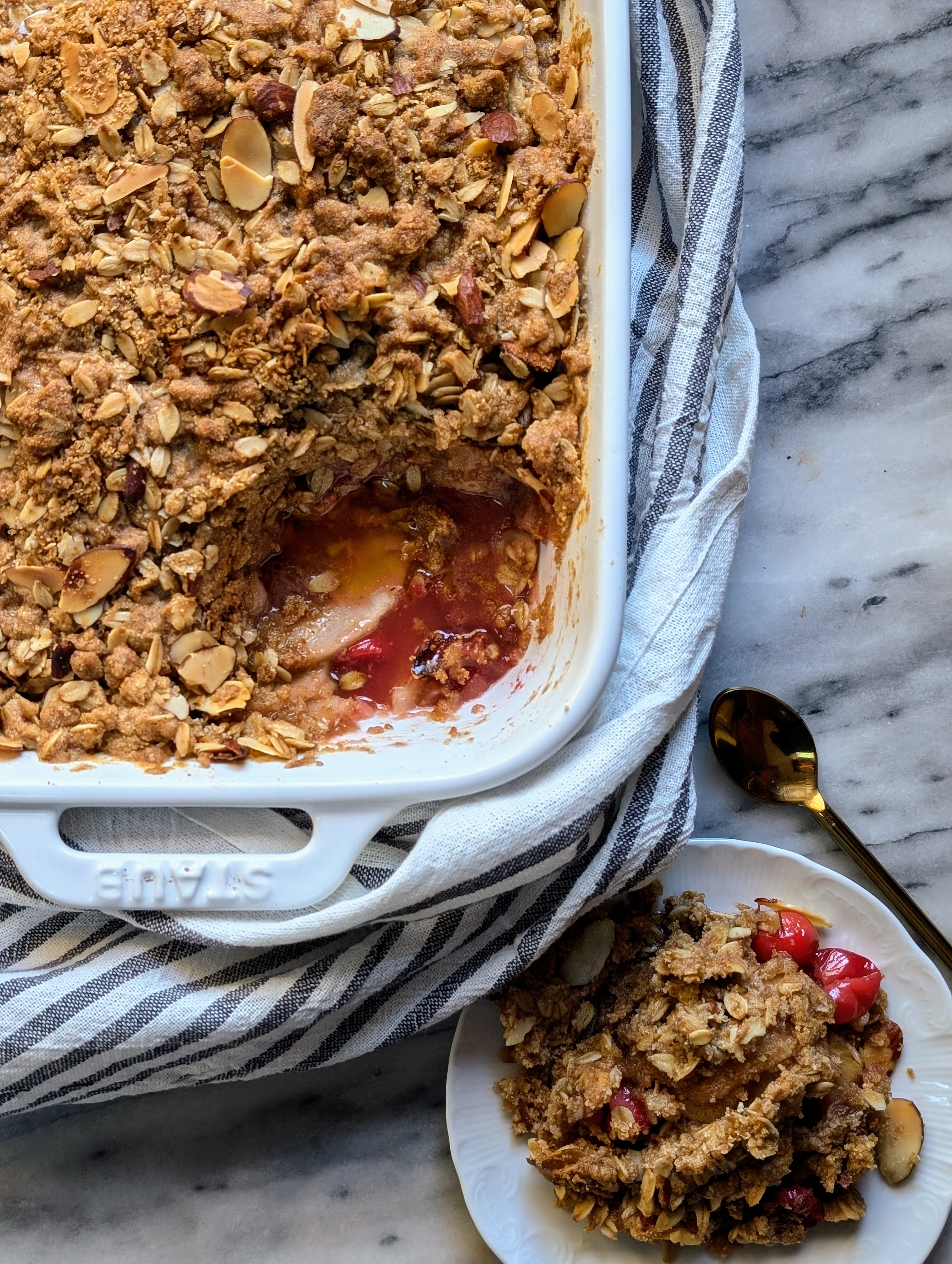 cranberry apple crumble in white baking dish nestled in striped dish towel. small plate of crumble next to dish with gold spoon.